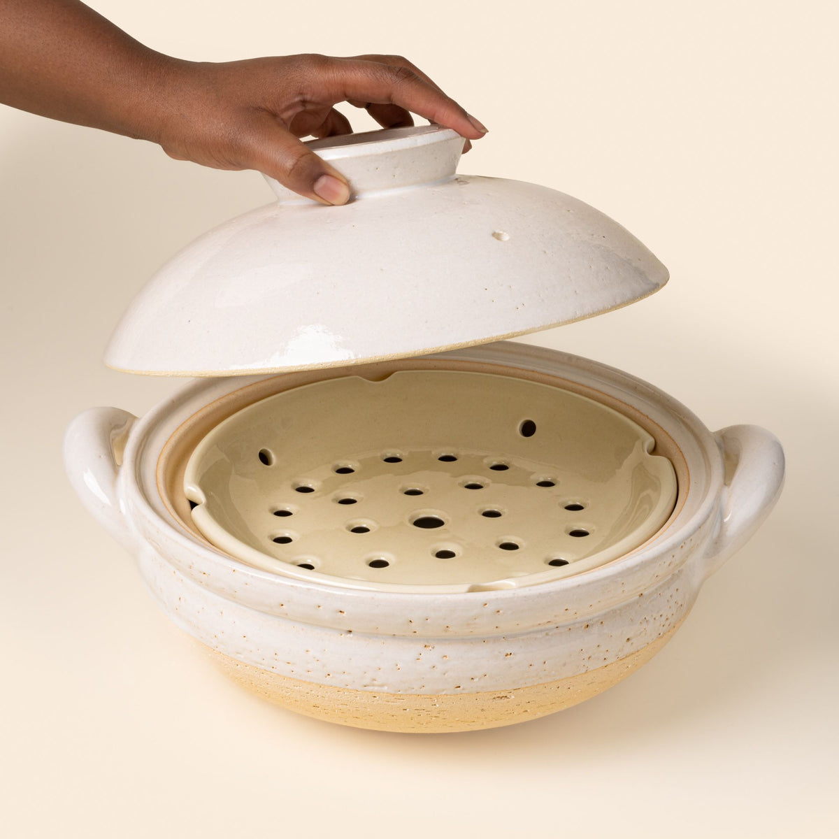 A hand holds a lid open above a white ceramic donabe steamer with side handles and a steamer tray