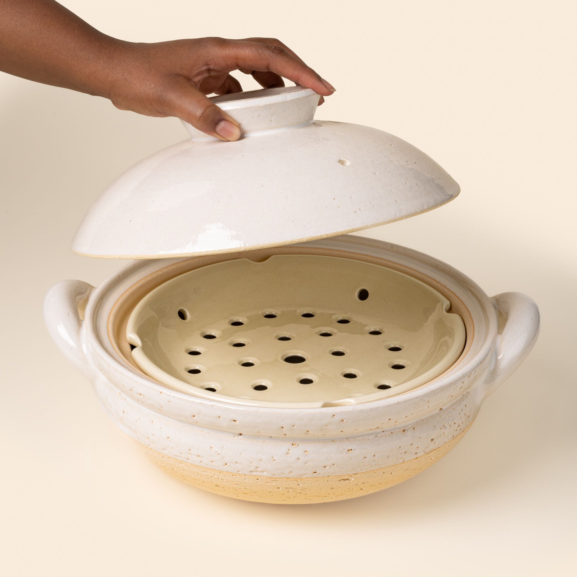 A hand holds a lid open above a white ceramic donabe steamer with side handles and a steamer tray
