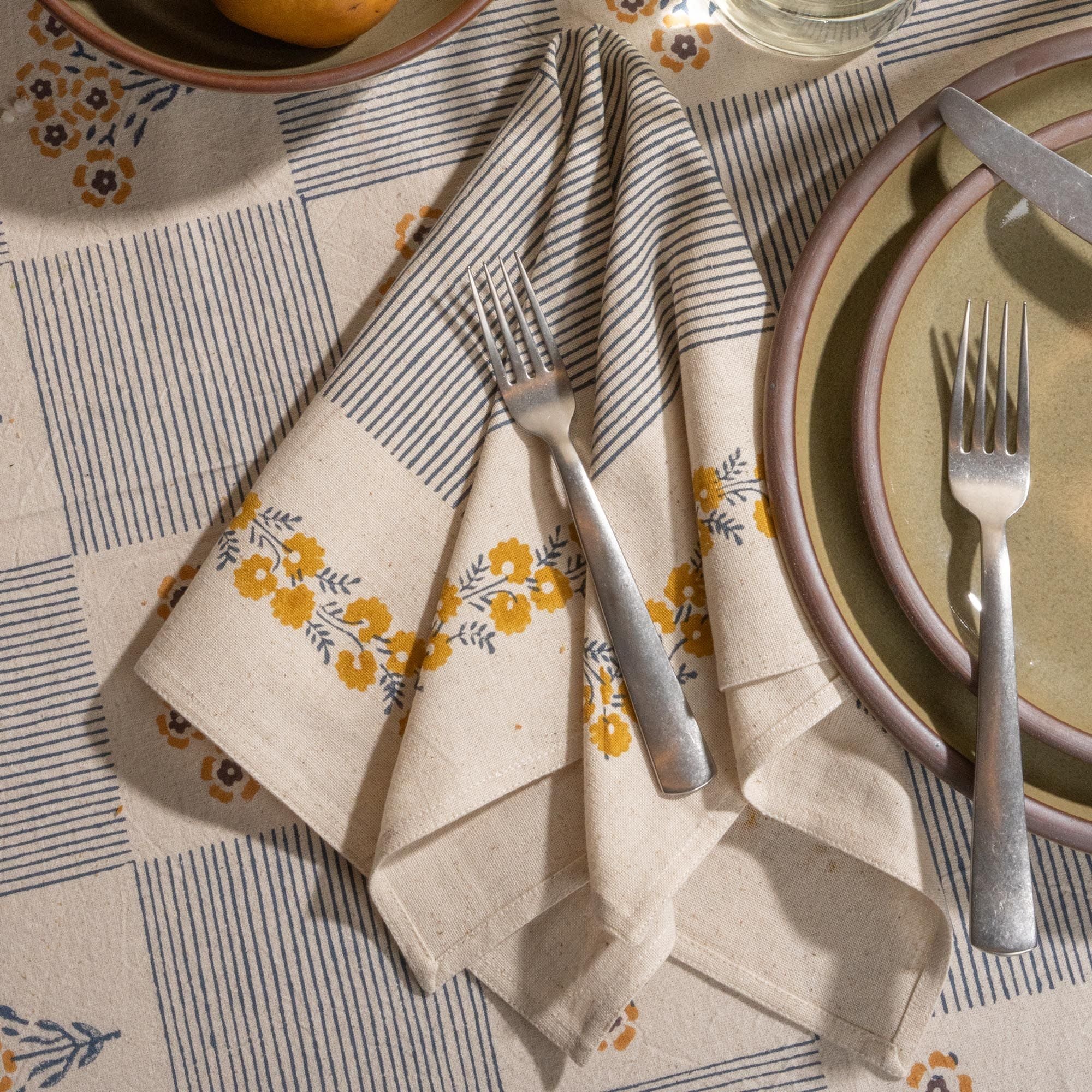 A cream-colored napkins with blue striped accents and yellow floral border detailing, arranged with silverware next to grey-green ceramic plates.