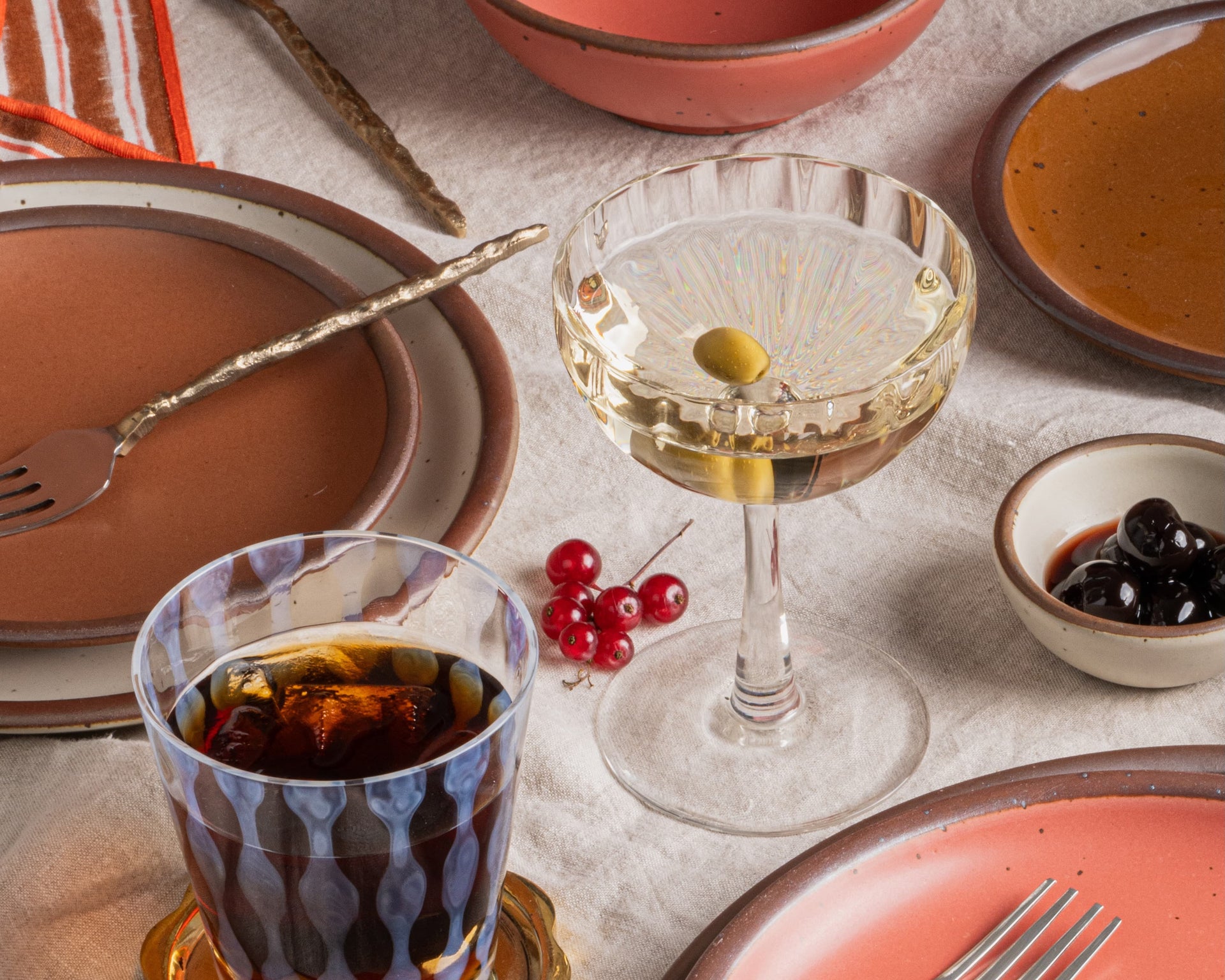 Styled tabletop scene with warm ceramic plates, vintage glassware, cocktails with olives, and small bowls of garnishes on a linen cloth.