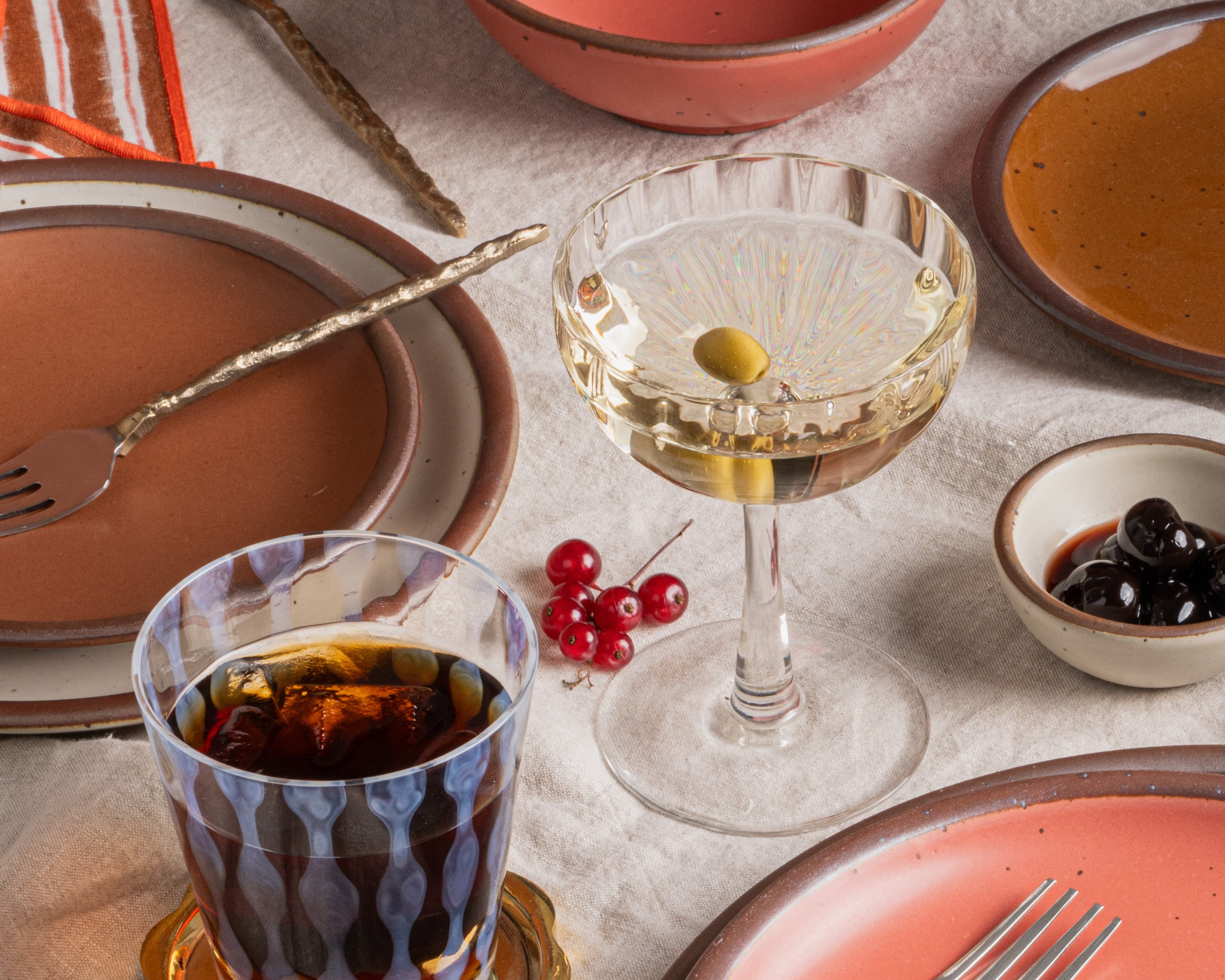 Styled tabletop scene with warm ceramic plates, vintage glassware, cocktails with olives, and small bowls of garnishes on a linen cloth.