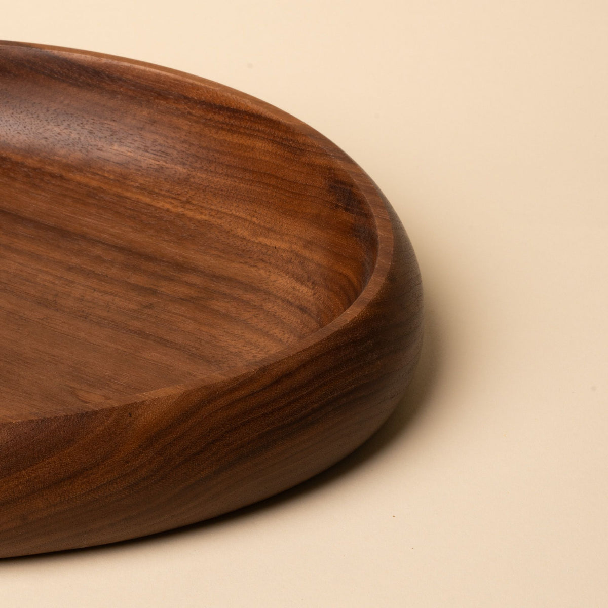 Close up of a shallow carved round walnut wood dish with smooth curved edges, resting on a beige background.