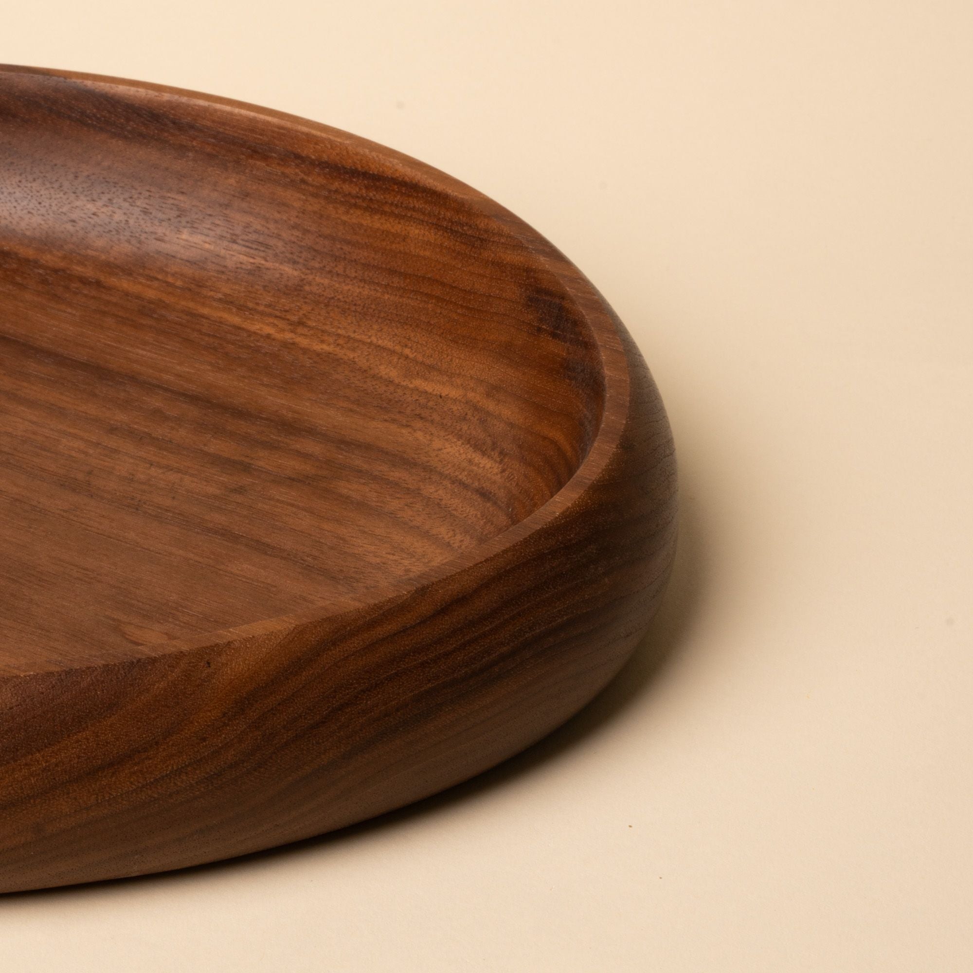 Close up of a shallow carved round walnut wood dish with smooth curved edges, resting on a beige background.
