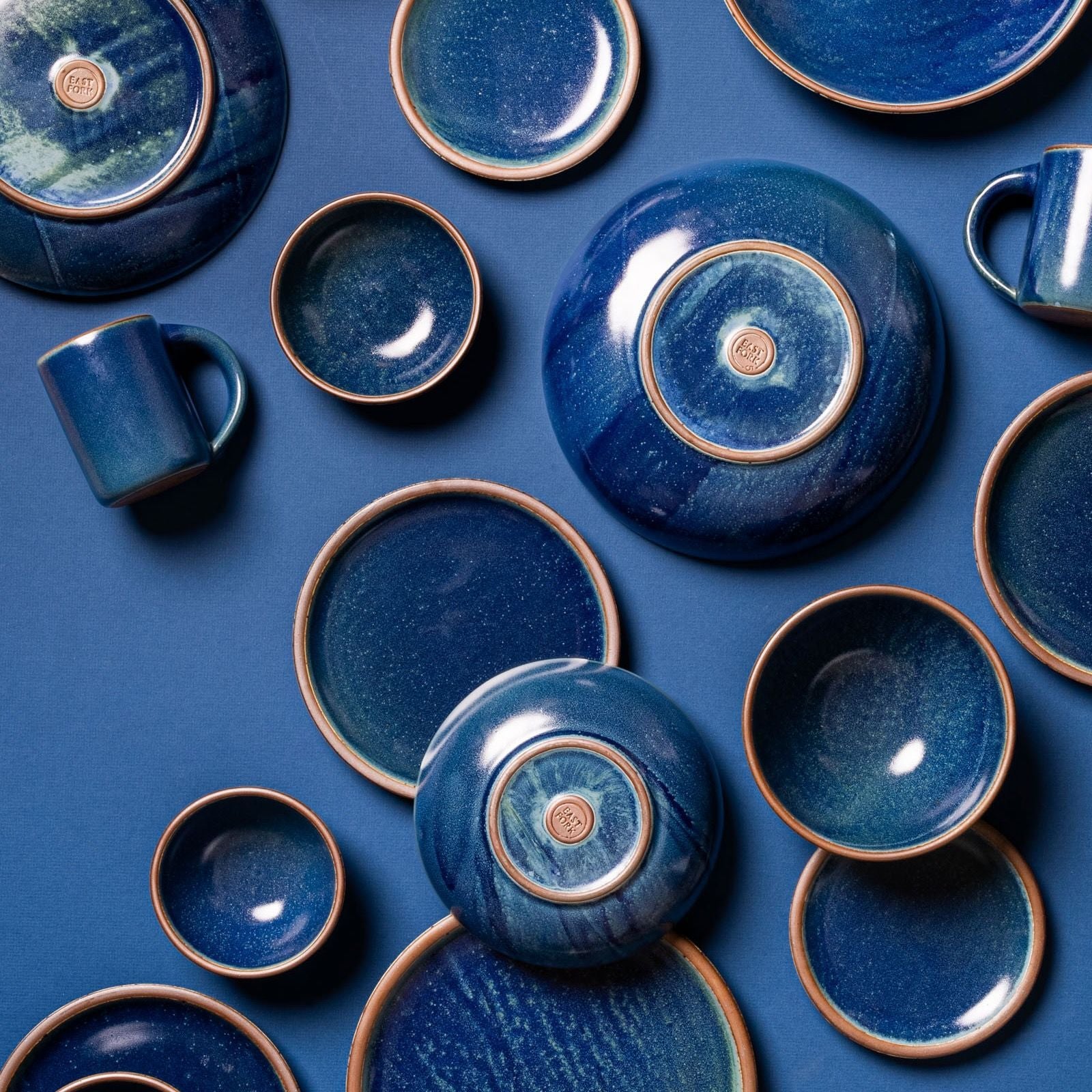 A collection of East Fork Pottery ceramics in a deep blue glaze, including plates, bowls, and mugs, arranged artistically on a matching blue background.