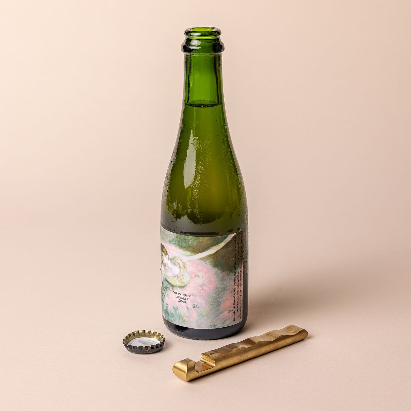 A green bottle of strawberry lavender cider with an artistic label stands on a beige background, accompanied by a twisted brass bottle opener and a removed bottle cap.