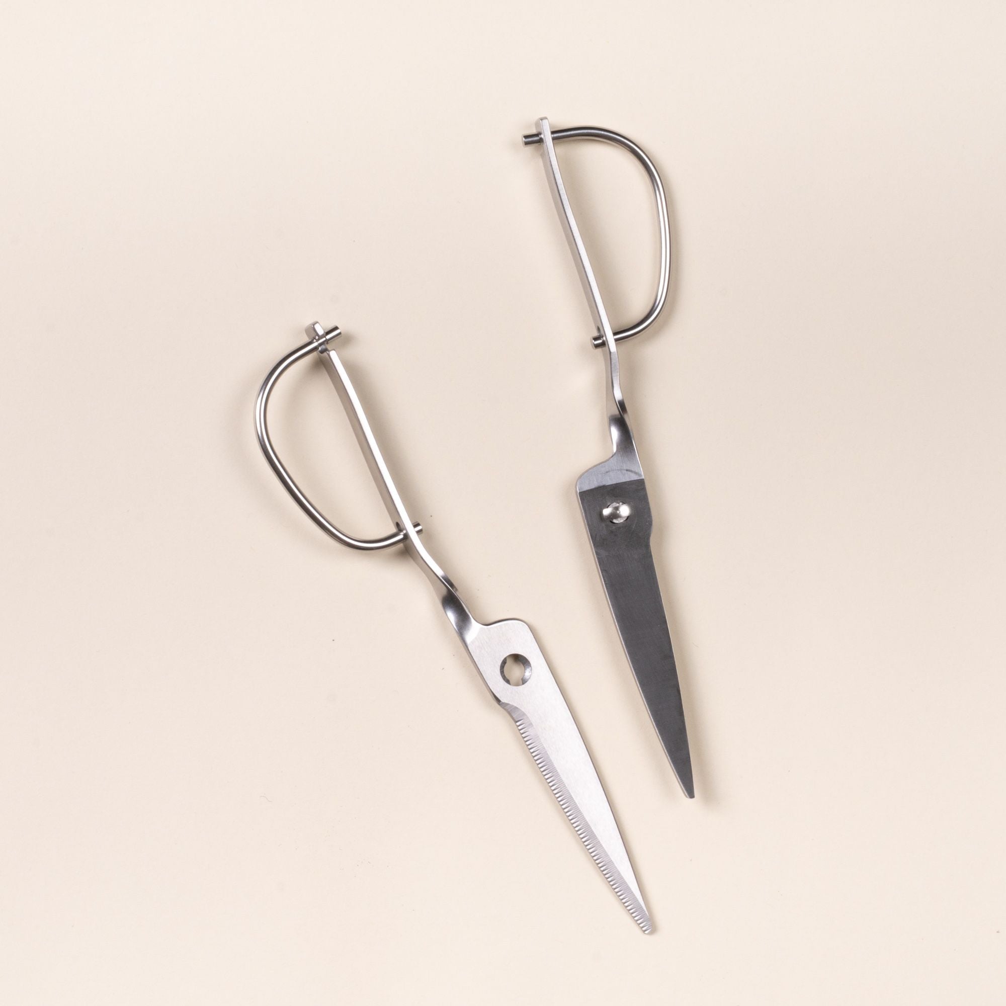 Two parts of stainless steel kitchen scissors with a looped handle, on a light neutral background.