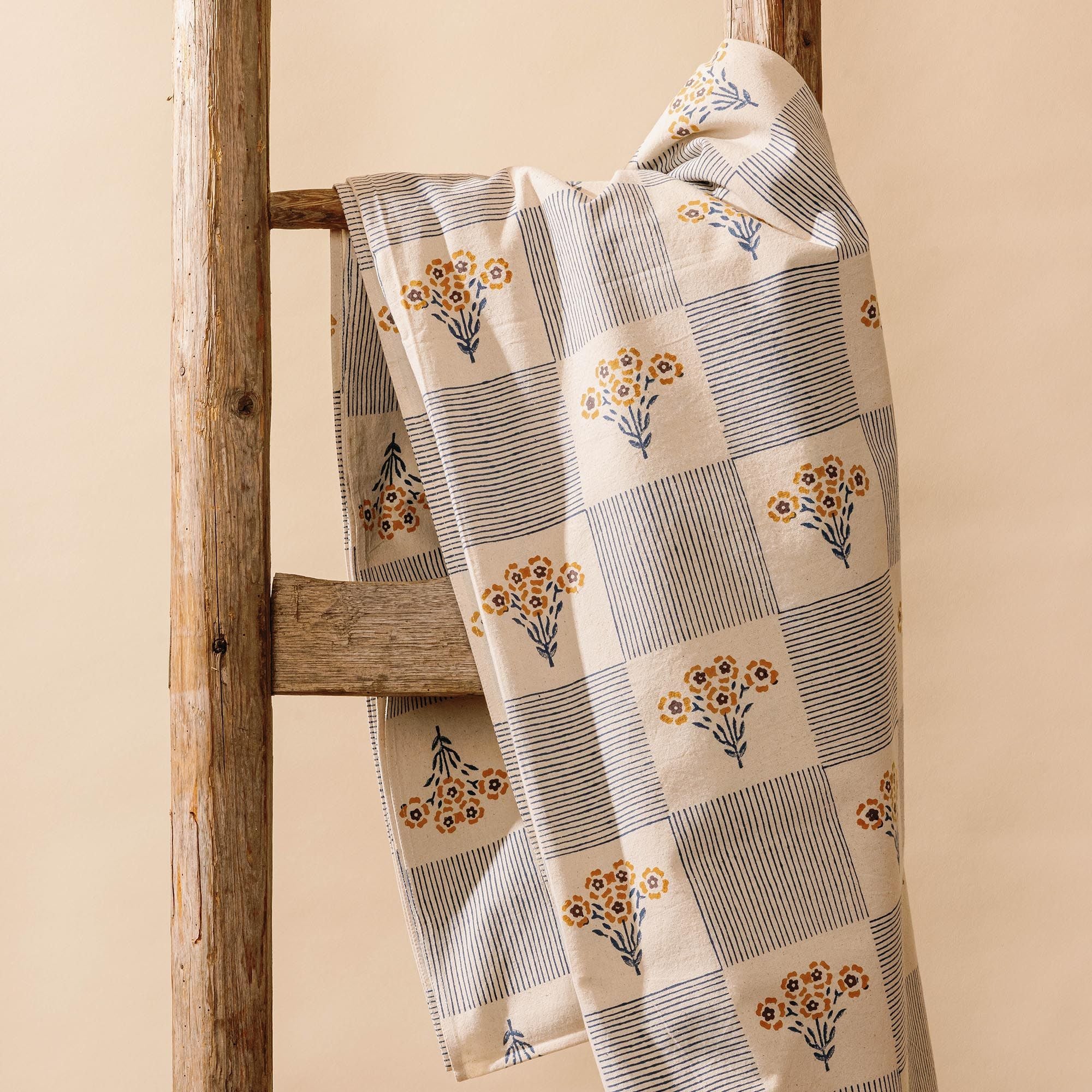 Folded blue-and-white checkered cloth tablecloth printed with small yellow flower bouquets on a wooden ladder.