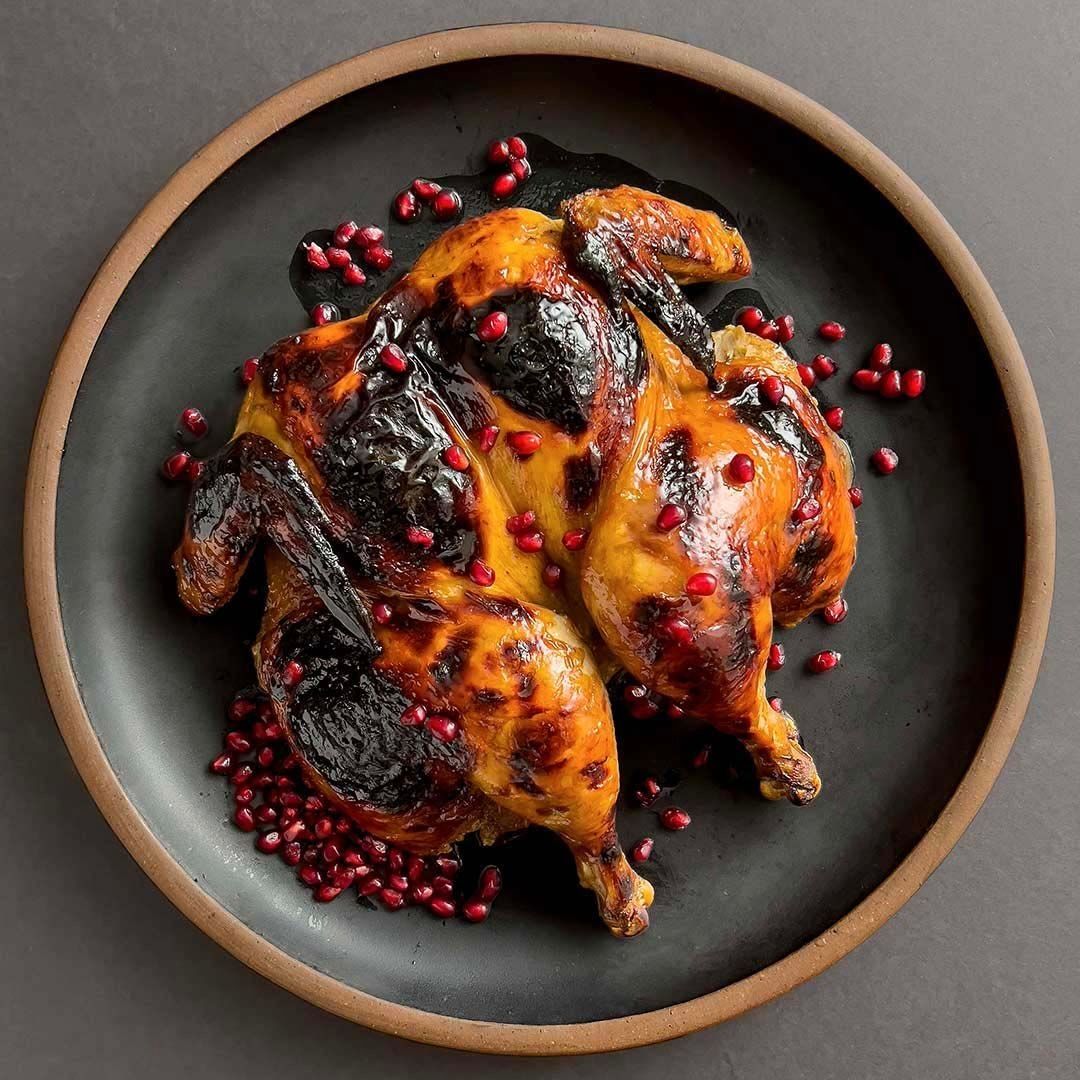 Roasted chicken with a charred exterior garnished with pomegranate seeds, presented on a dark ceramic plate with a brown rim.