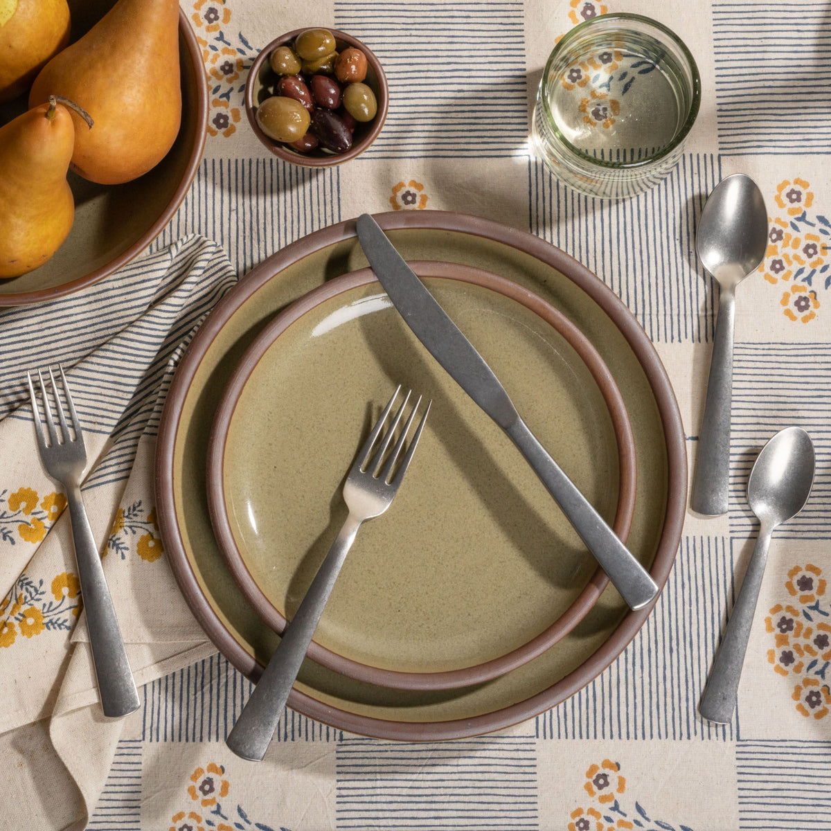 Place setting with reflective dappled grey-green plates, matte silver flatware, pears, olives, and water glass on a striped floral tablecloth.