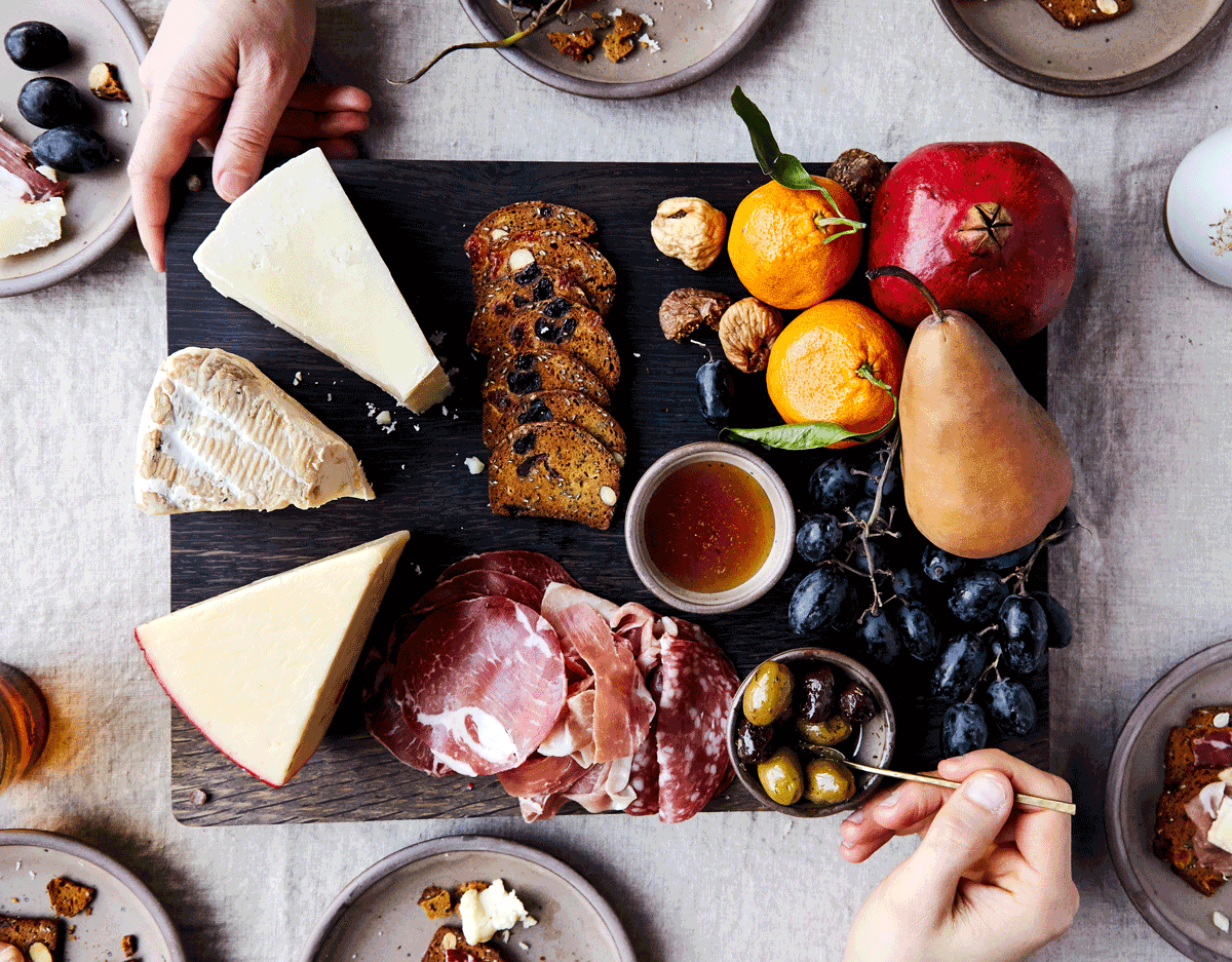 A charcuterie board on a grey tablecloth with various cheeses, sliced meats, fresh fruit, crackers, and a bowl of olives. Hands are reaching for items and placing food on surrounding plates.