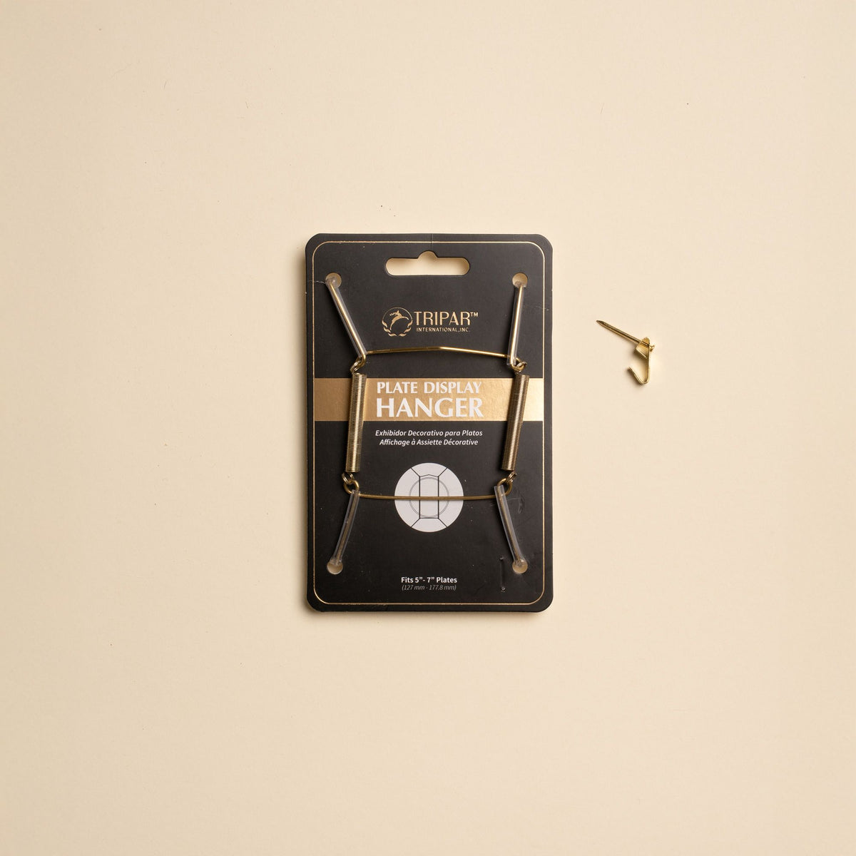 A brass plate hanger in its black cardboard package, with a nail off to the side.