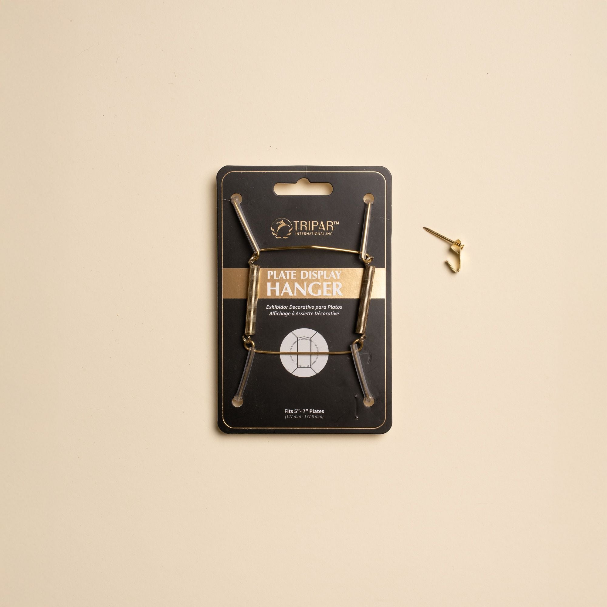 A brass plate hanger in its black cardboard package, with a nail off to the side.