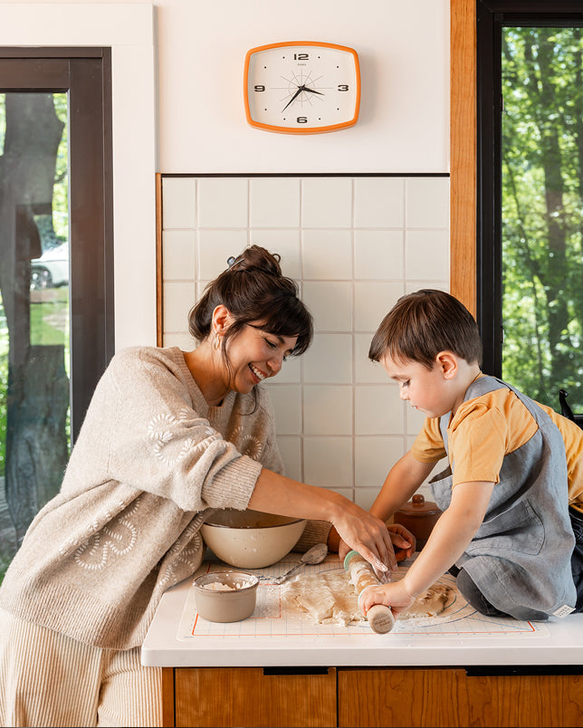 A woman and a young boy roll out dough on a white counter in a warm, modern kitchen. 