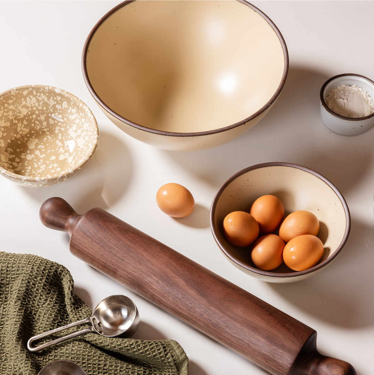 A styled flat-lay of baking and cooking tools on a light neutral surface, including a large cream-colored bowl with a unglazed rim, a smaller speckled bowl, a wooden rolling pin, metal measuring cups, a green cloth, and brown eggs in a cream bowl.