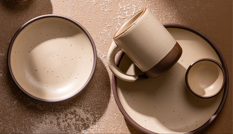A set of handmade ceramic tableware in a warm cream color with unglazed base including a bowl, a mug resting on a plate, and a small bowl, arranged on a textured brown surface.