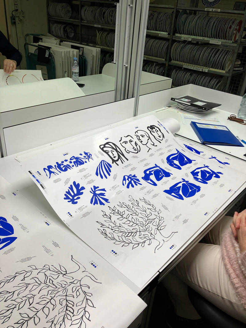 A person sits at a table examining large sheets of paper featuring blue and black artistic sketches, including abstract designs and faces. In the background, shelves hold stacks of plates, and various office supplies are on the table.