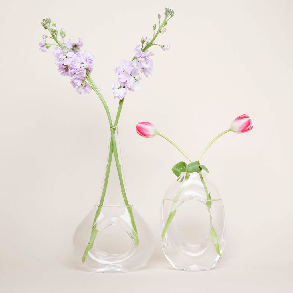 Two clear glass vases, one taller that the other, which have a hole at their center, creating a loop of glass, with two stems of flowers in each vase
