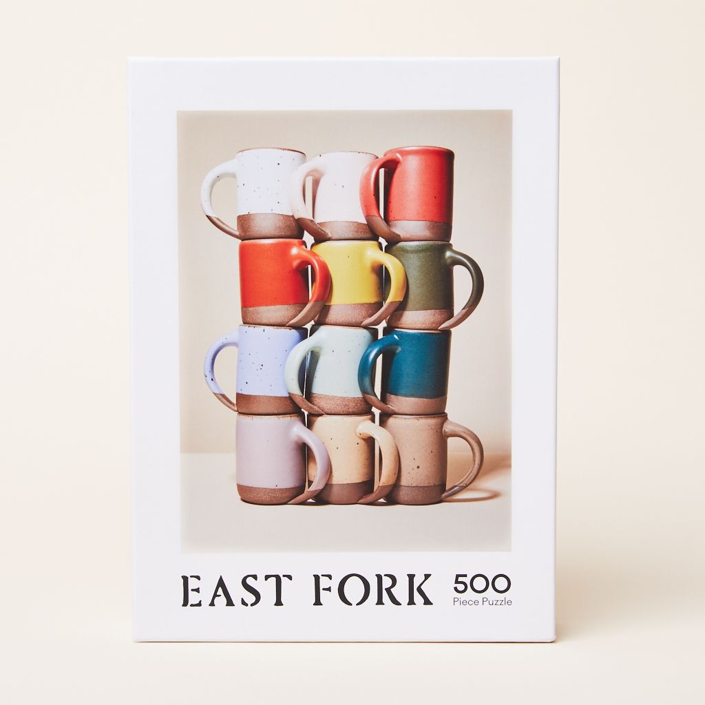 The front of the box of a 500-piece jigsaw puzzle that when put together shows 12 East Fork Mugs in 12 different glazes, stacked four high and three wide