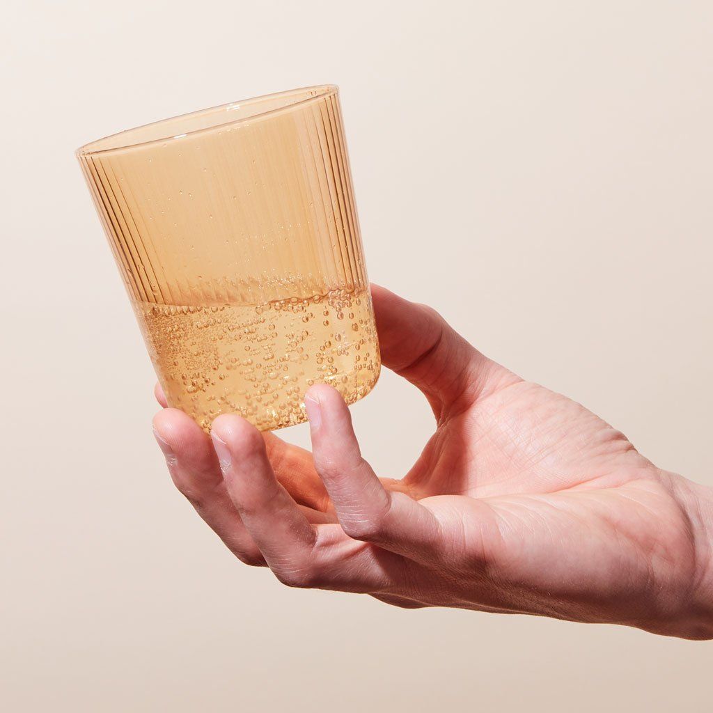A hand holds the base of a clear pale yellow drinking glass that is halfway full of a clear, bubbly liquid.