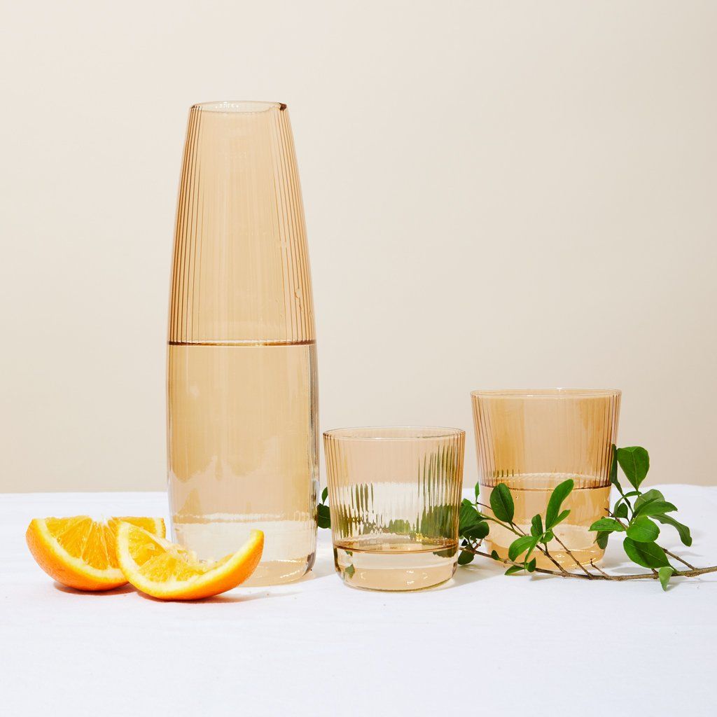 Wedges of oranges and a spring of herbs sit next to two pale yellow drinking glasses with matching pitcher with a spout at the rim