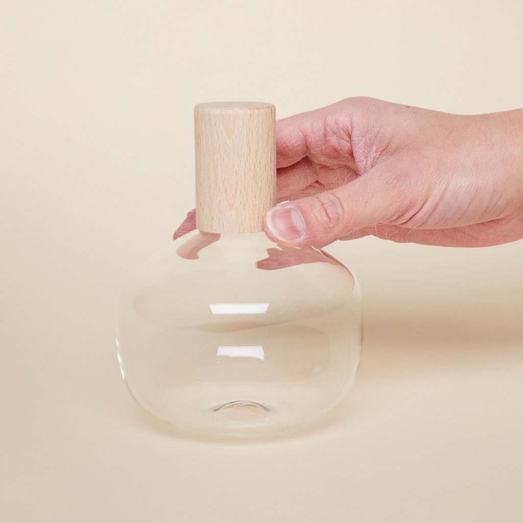 A hand holds a short glass bottle with wide body and a cylindrical wooden top