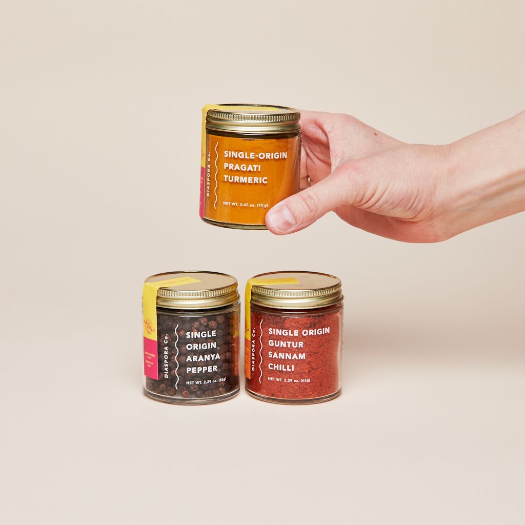 A hand holds a glass jar of turmeric above two jars, one holding pepper and the other, chillies