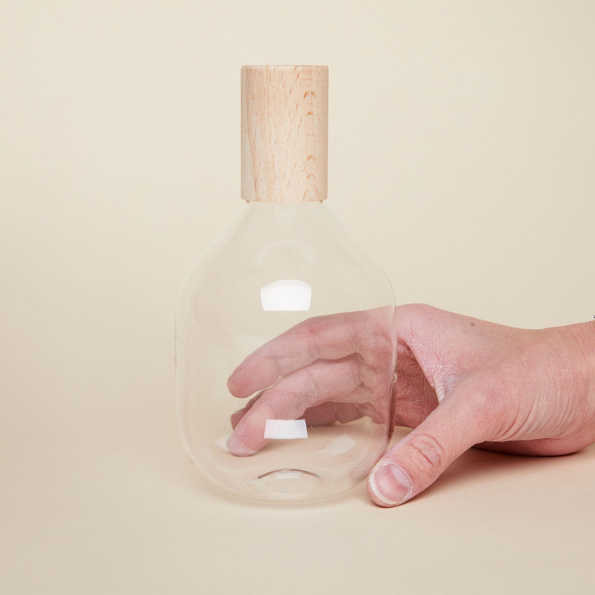 A hand holds a tall glass bottle with wide body and a cylindrical wooden top