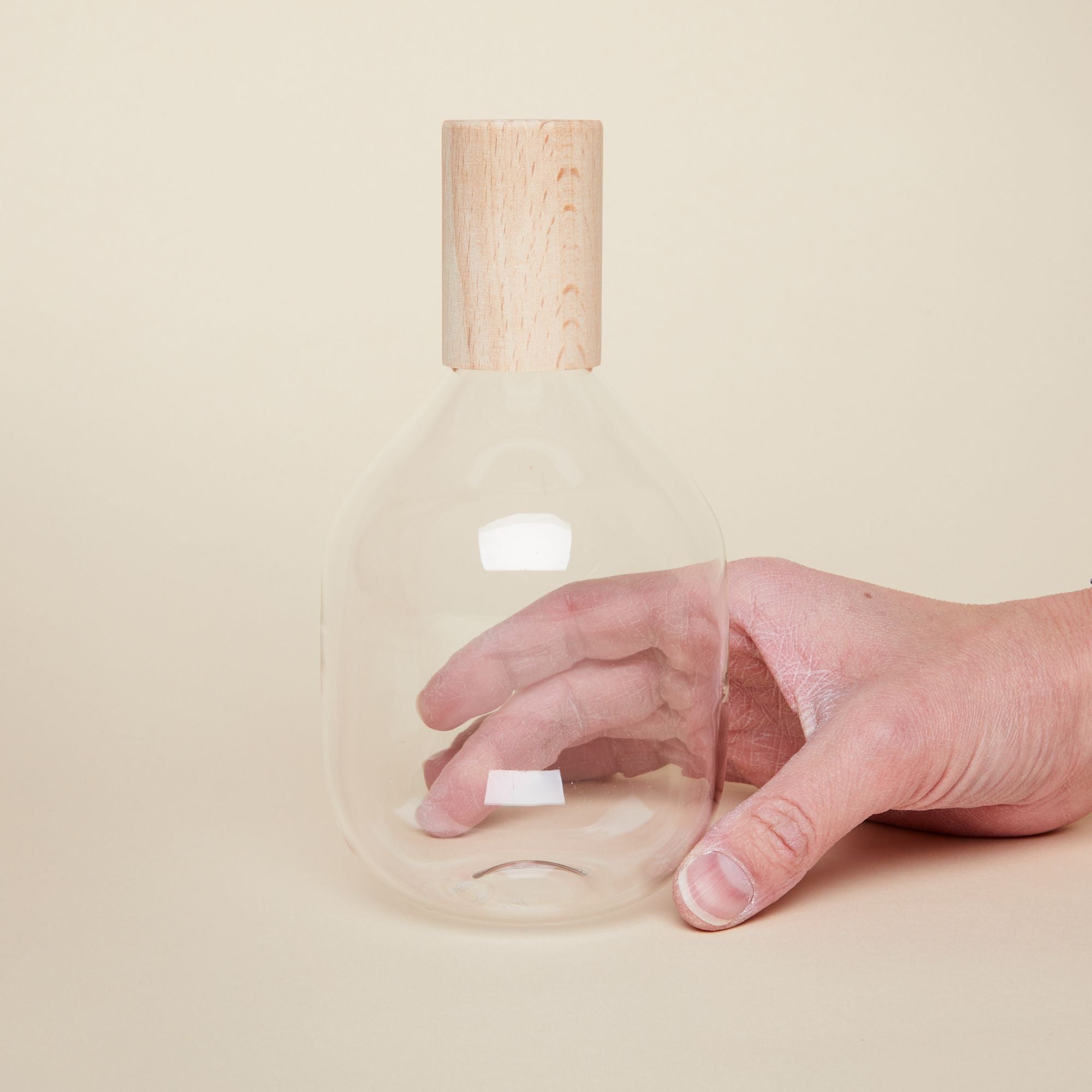 A hand holds a tall glass bottle with wide body and a cylindrical wooden top