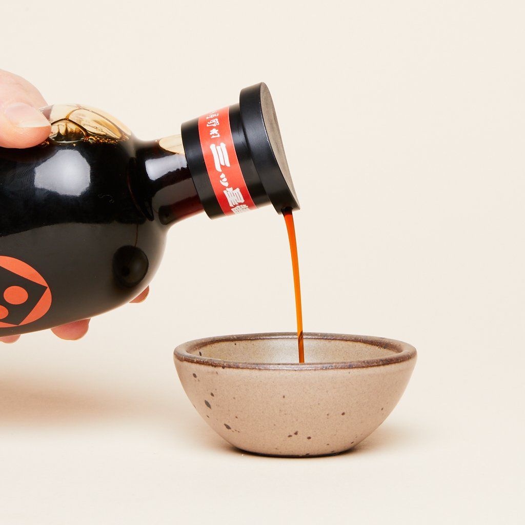 A hand pours a dark brown stream of soy sauce from its glass bottle into a Morel Bitty Bowl
