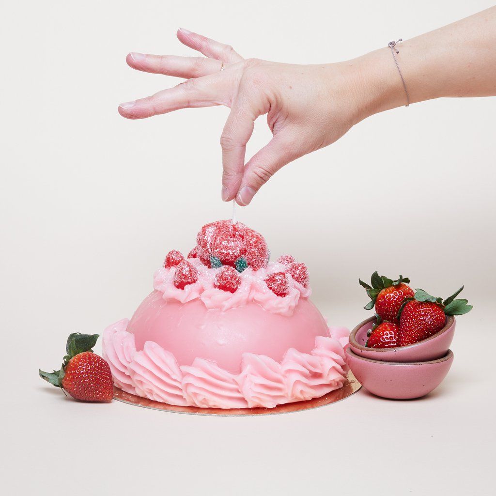 A thumb and index finger pinch the wick on top of a  dome-shaped pink candle that looks like a cake with icing and gumdrop flowers