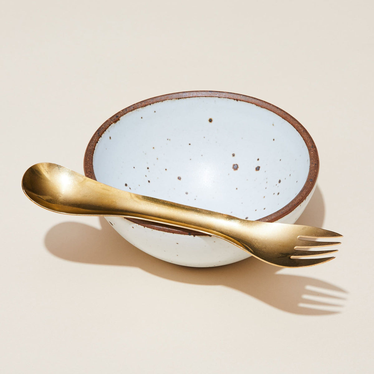 A brass eating utensil with spoon at one end and fork at the other, balanced atop an East Fork Breakfast Bowl